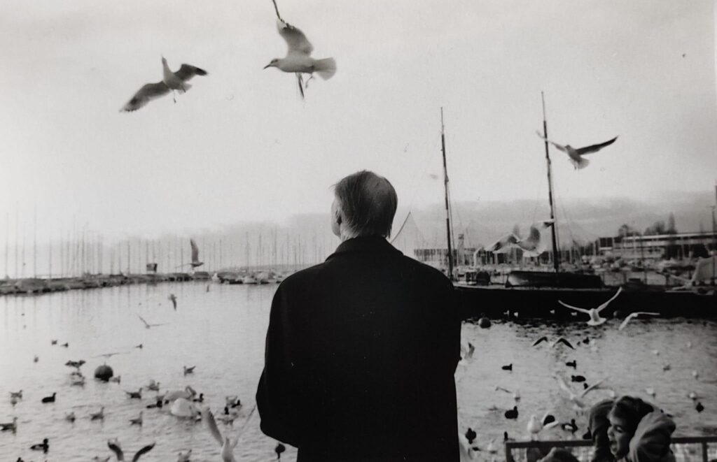 Luciano D'Alessandro - Napoli foto in bianco e nero di uomo di spalle davanti al mare con sullo sfondo un porto e molti gabbiani in acqua e in cielo