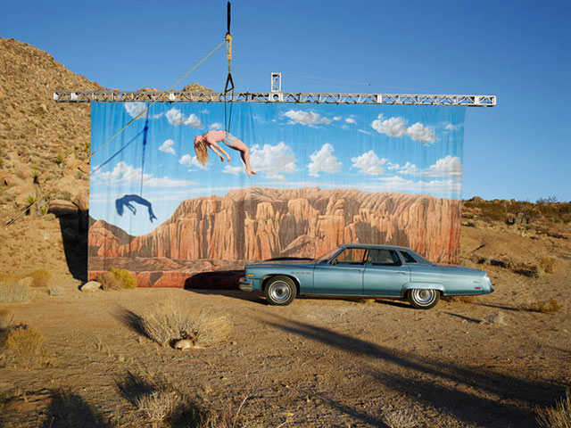 Sam Taylor-Johnson Roma foto a colori di macchina azzurra parcheggiata e donna appesa ad un cavo davanti a telo raffigurante paesaggio montano con cielo azzurro e nuvole