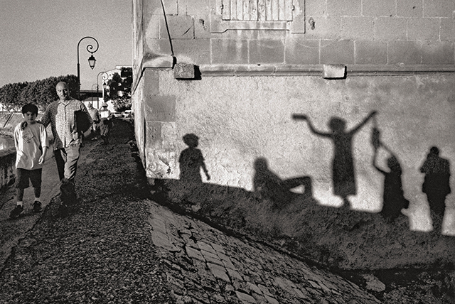 Gianni Berengo Gardin Brescia foto in bianco e nero di uomo e bambino che camminano per strada accanto a muro con riflesse delle ombre di altri bambini che giocano