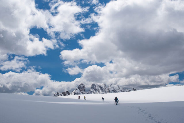 Sara Furlanetto Milano foto a colori di quattro alpinisti che camminano nella neve fra le montagne con cielo azzurro pieno di nuvole