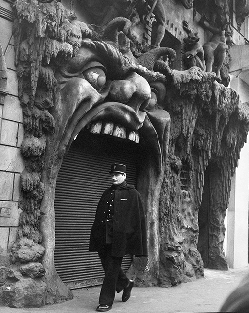 Robert Doisneau Milano foto in bianco e nero di poliziotto che cammina per strada davanti a negozio con insegna di mostro con enorme bocca aperta