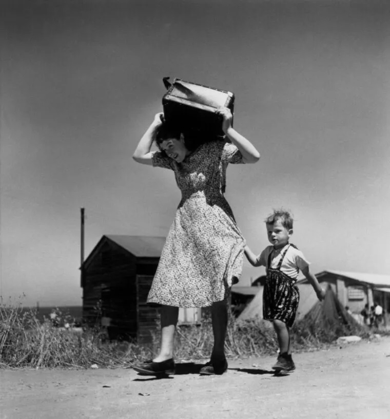 Robert Capa Aosta foto in bianco e nero di donna che cammina per la strada con pacco sulle spalle e bambino che le tiene la gonna