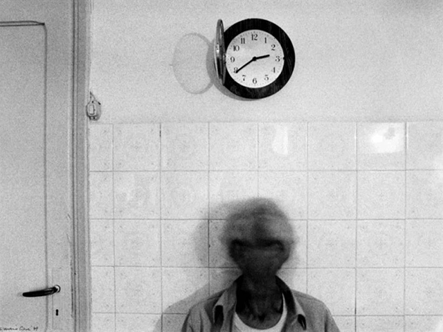 Mario Cresci Roma foto in bianco e nero di figura di uomo mossa appoggiato al muro di mattonelle bianche con orologio sopra la testa