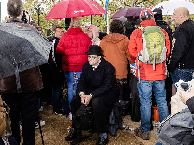 Guido Calamosca Senigallia foto a colori di uomo vestito di nero seduto su una sedia alle spalle di gruppo di persone con ombrello