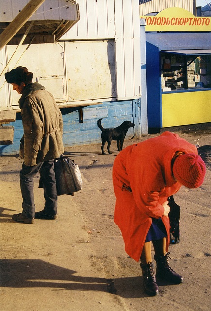 Boris Mikhailov Roma foto a colori di donna per strada con cappotto arancione che di tocca ginocchio e uomo di spalle con borsa in mano